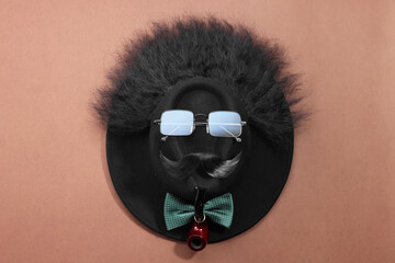 Man's face made of artificial mustache, sunglasses and hat on brown background, top view