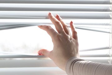 Woman separating slats of white blinds indoors, closeup