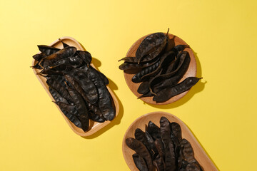 Different shape of wooden tray contains black locust fruits against on yellow background. High angle view with blank space for displaying product or adding text