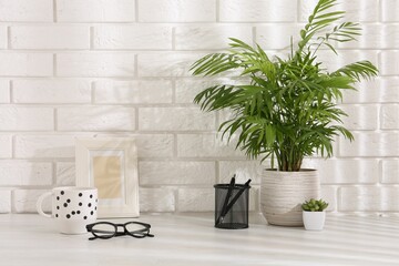 Stylish office workplace. Frame, plants, glasses, pens and cup on white table near brick wall