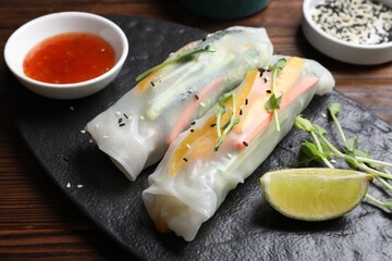 Tasty spring rolls, sauce, lime, sesame seeds and microgreens on wooden table, closeup