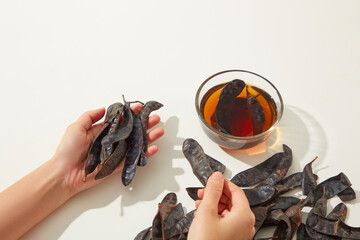 Top view scene that left hand is holding some black locust pods and right hand is lifting others one, a glass bowl of locust liquid placed on white table. High angle shot, copy space