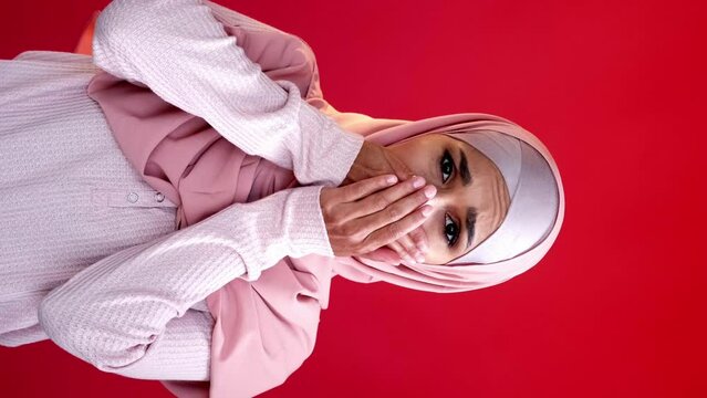 Vertical video. Omg shock. Regret sorrow. Grief compassion. Overwhelmed sad upset woman in hijab covering mouth with hands isolated on red copy space background.