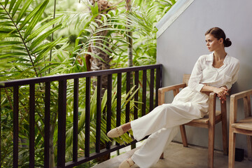 Smiling woman enjoying tropical vacation on balcony, sipping a drink in trendy sunglasses...