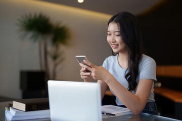 Asian teenage student woman using smartphone to chatting with friends and doing assignment homework after watching lecture video online lesson on laptop and studying about education knowledge in cafe