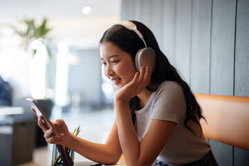 Asian teenage student woman wearing headphone to listening music and surfing social media on smartphone for relaxation after studying and learning education knowledge for preparing test exam in cafe