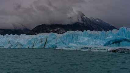 A wall of blue ice rises above a glacial turquoise lake. Thawed ice floes, icebergs floating in the...