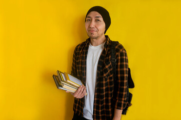 Portrait of young Asian student wearing beanie hat and carrying backpack, holds book while looking...