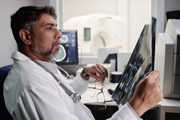 Side view of biracial male radiographer sitting at table in office working with head X-ray scan image - Powered by Adobe
