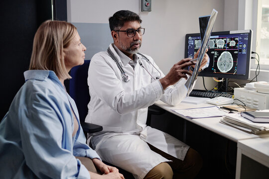 Biracial radiographer showing brain X-ray image to mature Caucasian female patient after CT procedure, copy space