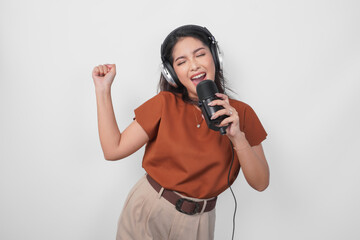 Excited young woman wearing brown shirt and headphone to listen music and singing along to the microphone isolated over white background.