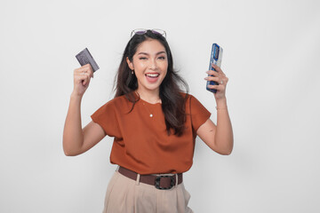 Happy successful Asian woman wearing brown shirt is holding her smartphone and credit card over isolated white background.