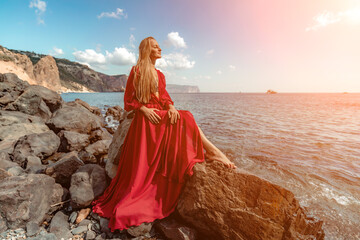 Red dress sea woman. A blonde with flowing hair in a long red dress sits on a rock near the sea....