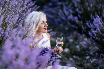 Blonde lavender field holds a glass of white wine in her hands. Happy woman in white dress enjoys...