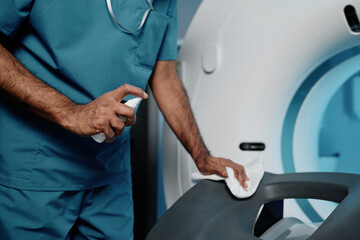 Unrecognizable male radiographer cleaning CT scanner bed with detergent at work in modern hospital,...
