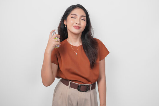 Beautiful young Asian woman in brown shirt holding bottle of perfume and spraying on her neck isolated by white background.