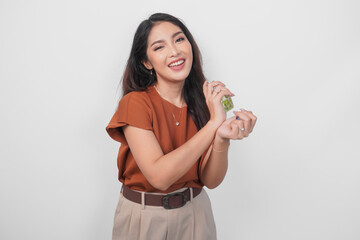 Smiling young Asian woman in brown shirt holding bottle of perfume and spraying on her wrist...
