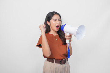 Happy successful Asian woman wearing a brown shirt raising clenched fist while shouting and screaming loud using megaphone speaker. Communication concept.