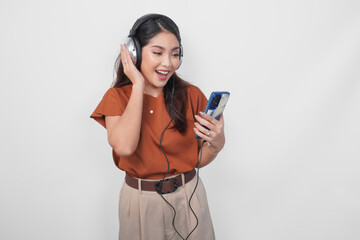 Cheerful young Asian woman in brown shirt listening to music from smartphone using wireless headphone.