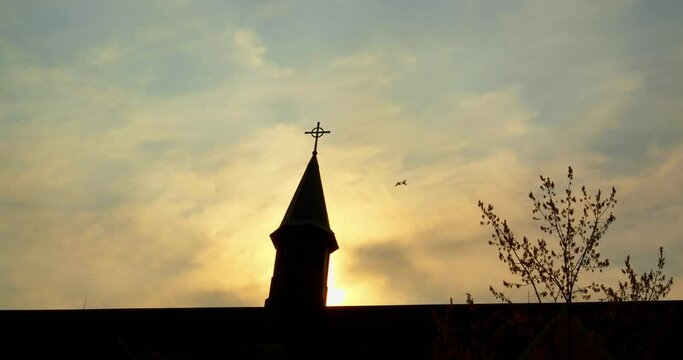 Birds at dusk, circling Catholic church spire, manifest as faith messengers against serene sunset backdrop. cross atop a Catholic church silhouetted by twilight, with birds embodying spirit of faith