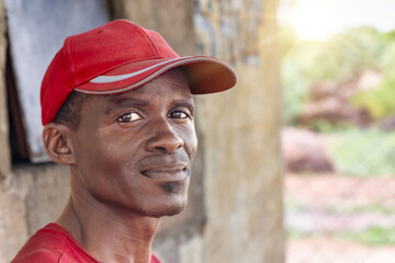happy village african man, standing outdoors in front of the house in the yard at sunset, he is wearing a red cap