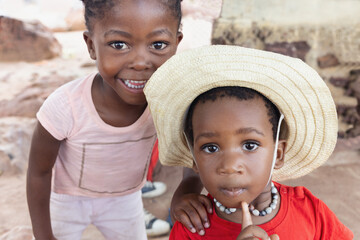 two small african kids in front of the house in the yard, wearing a red t-shirt and a straw hat the other with braids