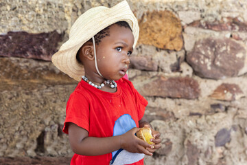 hungry small village african girl eating biscuits in front of the house in the yard, wearing a red t-shirt and a straw hat