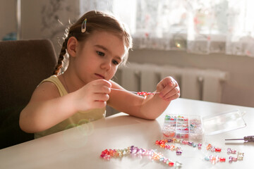 A young girl with pigtails creates colorful jewelry by stringing beads at a well-lit table, surrounded by an assortment of beads and string, showcasing her joy and focus in crafting. - 794787969