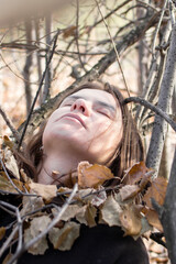 A young woman lies on a bed of autumn leaves and branches, her eyes closed and her face serene. She is lost in thought, perhaps about the beauty of the season or the changes that are to come.