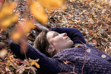 A serene scene of a young woman lying amongst autumn leaves, eyes closed in tranquility. The high-angle shot captures her peaceful expression amidst the brown and yellow leaf-covered ground.