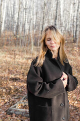 A young woman wearing a black coat stands pensively in a sparse autumn forest. The trees are bare, and the leaves are scattered on the ground. She looks off into the distance, lost in thought.