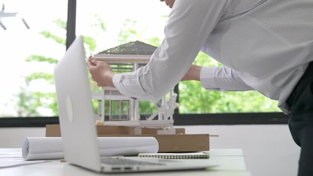 Close up of professional male architect engineer measure house model and using laptop analysis data at meeting table with house model and map . scatter around. Focus on hand. Immaculate.