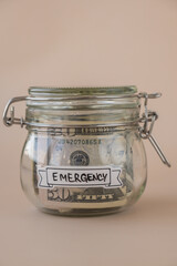 Saving Money In Glass Jar filled with Dollars banknotes. EMERGENCY transcription in front of jar. Managing personal finances extra income for future insecurity. Beige background
