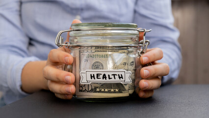 Female hands Saving Money In Glass Jar filled with Dollars banknotes. HEALTH transcription in front of jar. Managing personal finances extra income for future insecurity background