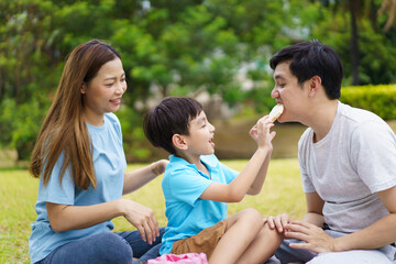 Happy cheerful Asian family with father, mother, and little son enjoy picnic together in a weekend at a park. Happy family moment concept. Parent and their child relax together in the park.