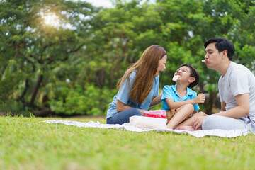 Fototapeta premium Happy cheerful Asian family with father, mother, and little son enjoy picnic together in a weekend at a park. Happy family moment concept. Parent and their child relax together in the park.