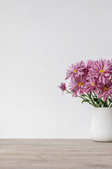 Empty white plaster wall, beige wooden table surface background, vase with pink daisy flowers...