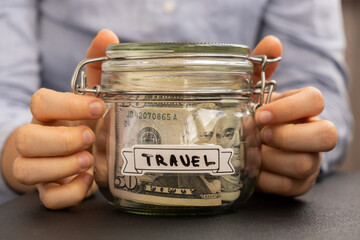 Unrecognizable woman holding Saving Money In Glass Jar filled with Dollars banknotes. TRAVEL transcription in front of jar. Managing personal finances extra income for future insecurity background