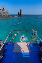 Tourist leaps from the back of a boat into clear blue ocean waters near a rocky formation