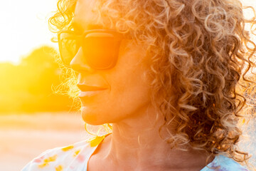 unny outdoor portrait of curly young woman smiling and wearing sunglasses with sunset backlight....