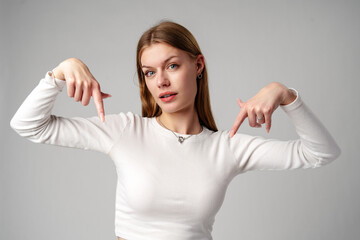 Young Woman in White Shirt Pointing Downwards With Both Hands Against a Gray Background