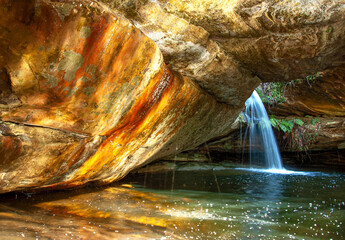 Beautiful orange color in rocks hanging over a pool of water with a waterfall and reflections.