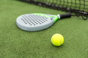 Yellow balls on grass turf near padel tennis racket behind net in green court outdoors with natural lighting. Paddle is a racquet game. Professional sport concept with copy space.