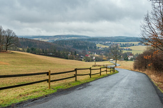 A winding country road that leads to a village where there are green hills with trees and cloudy skies all around.