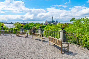 A peaceful view from King Oscar Terrace in Skansen showing wooden benches, lush trees, and distant city architecture under a bright blue sky. Stockholm, Sweden