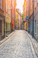 A narrow, sunlit cobblestone street lined with colorful historic buildings in the old town of Gamla Stan, Stockholm, Sweden, on a clear day.