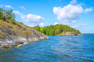 A vibrant view of the Stockholm Archipelago, showcasing lush greenery on rocky islands under a bright blue sky with scattered clouds. Sweden
