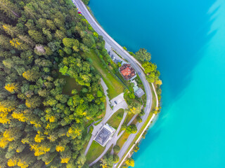 An aerial perspective showcases the vivid blue of a lake beside a road, contrasting with the rich green colors of the surrounding foliage and a solitary house nestled among the trees.