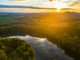 The sun sets behind a hilly landscape, casting a warm glow over the serene lake below, with trees lining its shores reflecting in the calm waters.