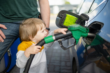 Cute little blond boy holding pump nozzle. Small funny kid helping father to fuel the car at a gas station. - obrazy, fototapety, plakaty
